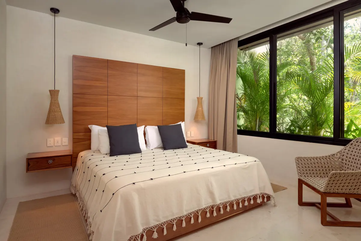 Casa Emilia bedroom with a tall wooden headboard, cream tasseled coverlet, woven pendant lamps and a window onto palms